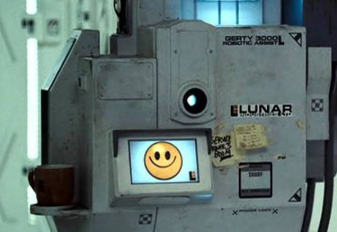 GERTY from the film Moon: a scuffed grey industrial robot with a single round camera lens, a LUNAR logo, handwritten notes taped to its side and a mug on a ledge, and a small screen showing a simple yellow smiley face
