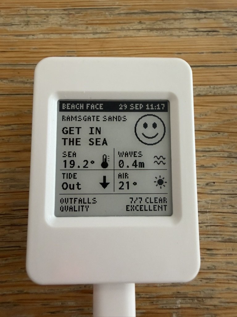 Beach Face on a wooden desk: a small white case with a square e-paper screen. BEACH FACE and the date along the top, RAMSGATE SANDS, GET IN THE SEA beside a big smiley face, then the sea at 19.2 degrees, waves 0.4m, tide out and air 21 degrees, each with a little icon, and 7/7 outfalls clear and excellent water quality along the bottom
